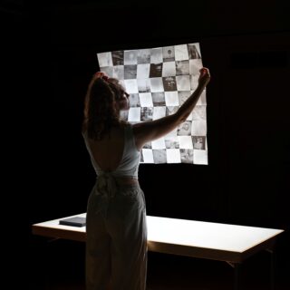 Eine Frau steht vor einem beleuchteten Tisch, hält ein großes Blatt mit Fotonegativen in der Hand, die in einem Schachbrettmuster angeordnet sind, und betrachtet es genau.
