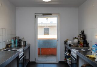 Kleine, schmale Küche mit zwei Elektroherden, Kochgeschirr, Geschirr und Reinigungsmitteln, mit Blick auf eine Glastür, die zu einem Außenbereich mit einer orangefarbenen Absperrung führt.