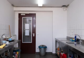 Eine kleine, schlicht ausgestattete Küche mit zwei Spülbecken, einer roten Tür, einem Mülleimer, Reinigungsmitteln und etwas Geschirr auf Arbeitsplatten aus Edelstahl unter Leuchtstoffröhren.