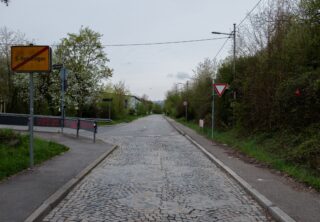 Eine leere Kopfsteinpflasterstraße mit einem Vorfahrtsschild auf der rechten Seite und einem gelben Ortsausgangsschild auf der linken Seite, gesäumt von Bäumen und unter bewölktem Himmel.