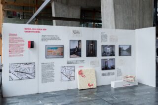 Ausstellungsaufbau mit Texttafeln, Karten, Fotos, Diagrammen und einem kleinen Architekturmodell, angebracht an einer weißen Wand in einem modernen Innenraum.