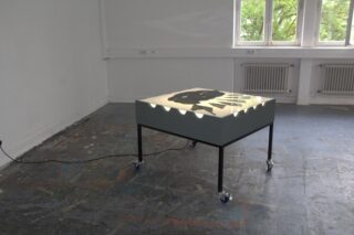 Ein Leuchtkasten mit einer schwarzen, abstrakten Form auf der Oberfläche steht auf Rollen in einem spärlich möblierten Raum mit großen Fenstern und einem abgenutzten Boden.
