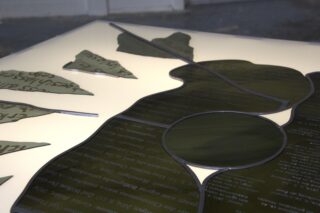 Beleuchtete Tafel mit durchscheinenden grünen Glasformen, auf denen Text eingraviert ist, vor einem leuchtend weißen Hintergrund.