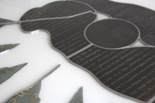 Metallkunstwerk mit eingraviertem Text und abstrakten Formen, die an Blätter und eine organische Gestalt erinnern, präsentiert auf einem weißen Untergrund.
