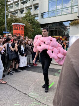 Eine Person, die im Freien in einer Menschenmenge spazieren geht und ein großes, rosa, wattiertes Kleidungsstück trägt, das wie abstrakte Blütenblätter geformt ist; im Hintergrund sind städtische Gebäude und Schaulustige zu sehen.