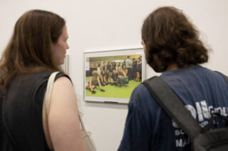 Zwei Menschen mit langen Haaren betrachten ein gerahmtes Foto an einer weißen Wand; das Foto zeigt eine Gruppe von Menschen, die gemeinsam in einem Raum auf einem grünen Boden posieren.