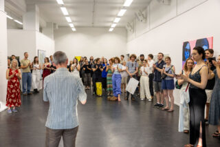Eine Gruppe von Menschen steht in einer Galerie mit weißen Wänden und blickt zu einem Mann, der gerade spricht. Viele klatschen, und an den Wänden sind farbenfrohe Kunstwerke zu sehen.
