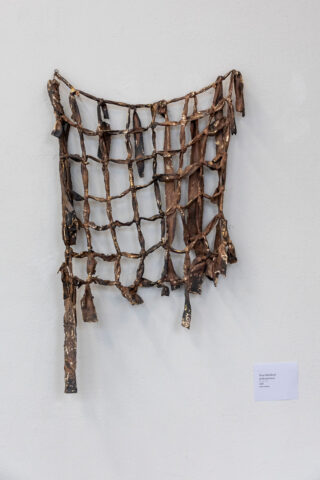 An einer weißen Wand hängt neben einem kleinen Schild eine handgefertigte, gitterartige Wandskulptur aus rauem, braunem und zerfetztem Material.