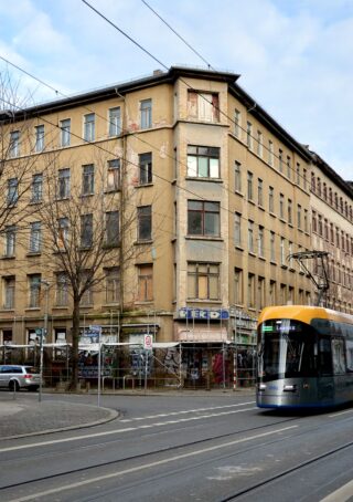 An einem bewölkten Tag fährt eine moderne Straßenbahn an einem alten, mit Graffiti übersäten Gebäude mit vernagelten Fenstern an einer Straßenkreuzung vorbei.