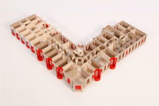 Ein V-förmiges Architekturmodell mit mehreren Räumen und roten Akzenten an den Ecken und Fenstern, von oben betrachtet vor einem einfarbigen Hintergrund.