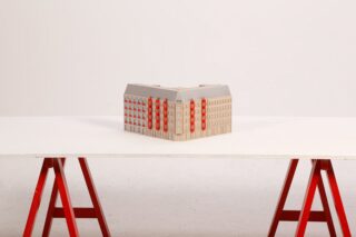 Ein kleines Architekturmodell eines beigen Gebäudes mit roten Akzenten steht auf einer weißen Tischplatte, die von roten Sägeböcken gestützt wird, vor einem schlichten weißen Hintergrund.