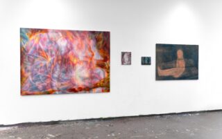 An einer weißen Wand in einer Kunstgalerie sind vier abstrakte Gemälde mit verschwommenen, farbenfrohen Formen ausgestellt. Auf dem Boden sind Farbflecken verstreut.