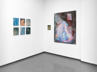 An einer minimalistisch gestalteten Galeriewand sind sechs kleine blaue abstrakte Bilder und zwei größere Gemälde mit wirbelnden Farben an einer weißen Wand über einem grauen Boden zu sehen.