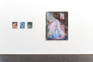 An einer weißen Galeriewand über einem dunklen Boden hängen vier abstrakte Gemälde, drei kleine und ein großes. Das große Gemälde zeigt verschwommene, farbenfrohe Formen und ein rotes, fensterähnliches Detail.