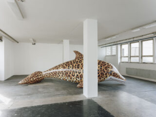 Eine große Skulptur eines Delfins mit Leopardenmuster ist in einem weißwandigen Raum im Industriestil mit Betonböden und Fenstern ausgestellt.