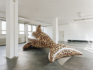 Ein großer aufblasbarer Hai mit Leopardenmuster steht auf dem Boden eines weißen Raums im Industrial-Stil mit Fenstern und freiliegenden Rohrleitungen.