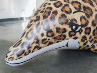 Ein großer aufblasbarer Delfin mit Leopardenmuster liegt auf einem Betonboden in einem hell beleuchteten Innenraum.