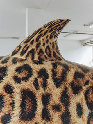Ein großes aufblasbares Objekt mit Leopardenmuster und einer Rückenflosse, das in einem Innenraum vor einem weißen Hintergrund platziert ist.