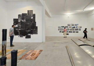 Drei Personen betrachten verschiedene Kunstwerke, darunter Fotos, Drucke und Bodeninstallationen, in einer geräumigen Galerie für zeitgenössische Kunst mit weißen Wänden.