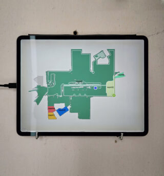 Auf einem Tablet wird ein digitaler Grundriss mit beschrifteten Bereichen angezeigt: Räume 1, 2, 3 und 4. Das Tablet ist über ein Kabel angeschlossen und an einer hellen Wand befestigt.