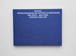 Auf einer Holzfläche liegt ein geschlossenes Buch mit dem Titel „OKUPO Verschiebungen im öffentlichen Raum Mai 2023 – Mai 2026 Janosch Müller“.