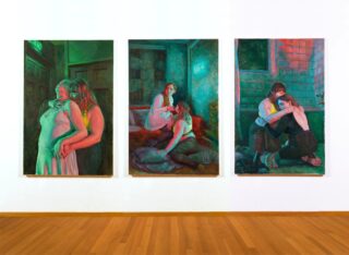 An einer weißen Wand hängen drei farbenfrohe Gemälde nebeneinander, auf denen jeweils zwei Frauen in emotionalen, intimen Posen in häuslichen Innenräumen dargestellt sind; der Boden besteht aus hellem Holz.