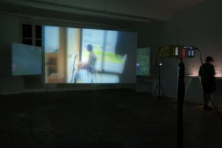 Ein schwach beleuchteter Raum mit einer großen, unscharfen Videoprojektion an der Wand, einem gelben Projektor im Vordergrund und einer Person, die neben einem kleinen beleuchteten Display steht.
