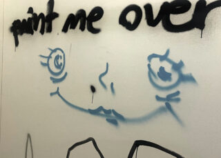 Graffiti an einer Wand, auf dem mit schwarzer Sprühfarbe „Paint me over“ über einem blauen, aus einfachen Linien gezeichneten Comic-Gesicht steht.