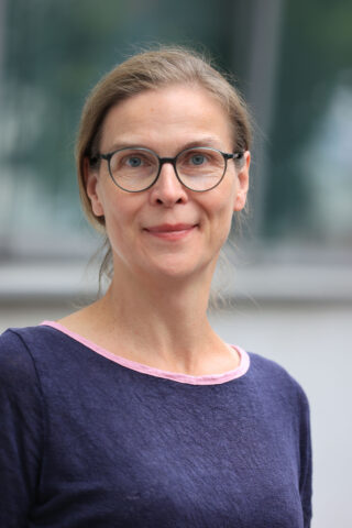 Eine Frau mit Brille und hellbraunen, nach hinten gestrichenen Haaren, die ein blaues Oberteil mit rosa Kragen trägt, steht vor einem unscharfen Hintergrund im Freien.