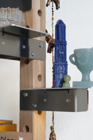 Auf einem Metallregal stehen eine blaue Turmfigur, ein lächelnder Pokal, ein kleines grünes Spielzeug und eine hängende Perlenverzierung vor einer hölzernen Lochwand.