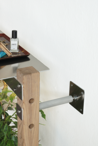 Eine Nahaufnahme eines Holzregals, das mit Metallhalterungen an der Wand befestigt ist und auf dessen unterer Ebene ein Tablett, eine kleine Flasche mit der Aufschrift „DIRTY“ sowie einige Pflanzen stehen.