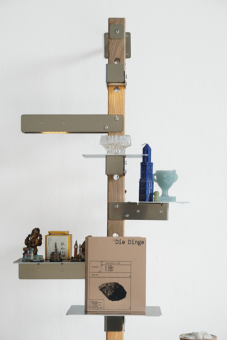 Auf einem vertikalen Holzregal mit Glas- und Metallstützen sind kleine Skulpturen, ein blaues Turmmodell, eine Zier-Tasse, Glaswaren und eine braune Mappe mit der Aufschrift „Die Dinge.“ ausgestellt.