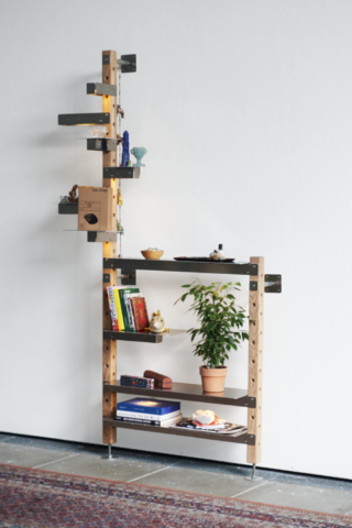 Ein Holzregal mit Metallhalterungen vor einer weißen Wand, auf dessen versetzt angeordneten Regalböden Bücher, eine Topfpflanze, Dekoartikel und kleine Gegenstände stehen.