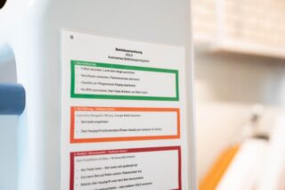 Nahaufnahme eines laminierten Beipackzettels mit drei farbcodierten Abschnitten und deutschem Text, der an einer weißen Maschine angebracht ist.