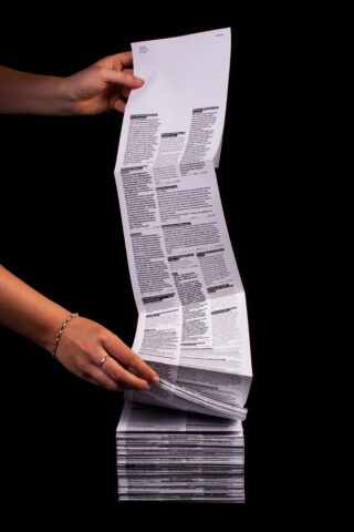 Zwei Hände halten ein langes, gefaltetes Blatt Papier mit dichtem Text fest und heben es an; es liegt auf einem Stapel ähnlicher Blätter vor einem schwarzen Hintergrund.