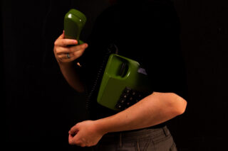Eine Person, die ein grünes Vintage-Schnurtelefon vor einem dunklen Hintergrund hält, wobei sie mit einer Hand den Hörer und mit der anderen die Telefonbasis festhält.