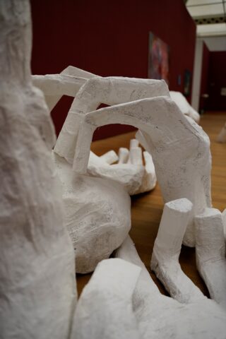 Weiße abstrakte Skulpturen, die an langgestreckte Hände und Finger erinnern, sind auf einem Holzboden in einer Kunstgalerie mit roten Wänden ausgestellt.