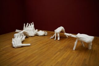 In einer Kunstgalerie sind weiße, abstrakte Skulpturen, die an Tier- und Baumformen erinnern, auf einem Holzboden vor einer dunkelroten Wand ausgestellt.