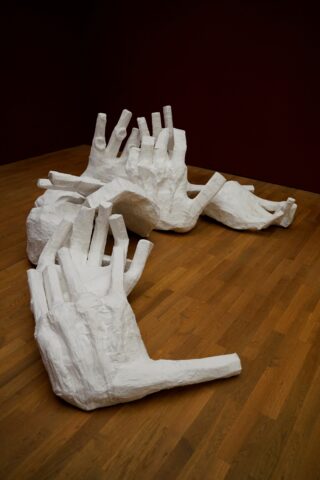 Große, weiße, skulpturale Hände aus Gips oder einem ähnlichen Material sind in einer wellenförmigen Anordnung auf dem Boden einer Holzgalerie vor einer dunkelroten Wand angeordnet.