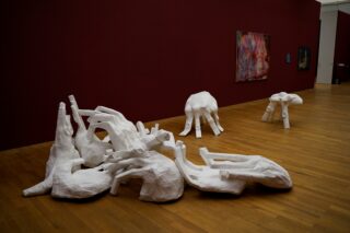 Eine Galerie für moderne Kunst mit abstrakten weißen Skulpturen auf einem Holzboden und farbenfrohen Gemälden an einer kastanienbraunen Wand.
