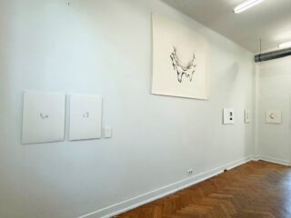 An einer minimalistisch gestalteten Kunstgalerie-Wand sind mehrere abstrakte Schwarz-Weiß-Skizzen auf weißem Hintergrund über einem Fischgrätenparkett zu sehen.