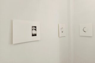 An einer weißen Galeriewand sind drei minimalistische Schwarz-Weiß-Zeichnungen angebracht, die jeweils mittig auf einzelnen rechteckigen Papierbögen mit breiten Rändern platziert sind.