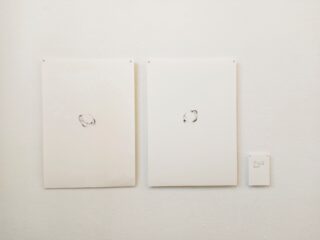 An einer weißen Wand sind zwei weiße Blätter mit kleinen, einfachen kreisförmigen Skizzen angeheftet; rechts davon ist ein kleines gedrucktes Etikett angebracht.