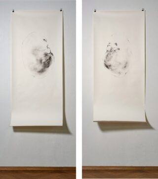 An einer Wand hängen nebeneinander zwei große Papierbögen, auf denen jeweils in der Mitte eine blasse, abstrakte Kohlezeichnung zu sehen ist, die einer wolkenartigen oder rauchartigen Form ähnelt.