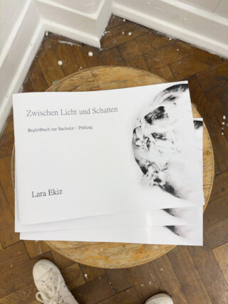 Auf einem Holzhocker liegt eine Broschüre mit dem Titel „Zwischen Licht und Schatten“ von Lara Ekiz; auf dem braunen Holzboden ist ein weißer Sneaker zu sehen.