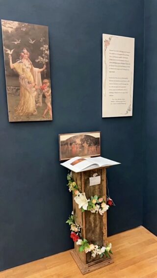 Eine Museumsausstellung zeigt ein Gemälde, eine Texttafel und ein aufgeschlagenes Buch auf einem mit Blumen geschmückten Holzsockel vor einer dunkelblauen Wand.