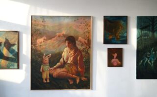 An einer weißen Wand hängen fünf surrealistische Gemälde, darunter ein großes Bild, auf dem eine Frau neben einem Kind sitzt, das eine Hasenmaske trägt, während im Hintergrund rosa Tiere und Berge zu sehen sind.
