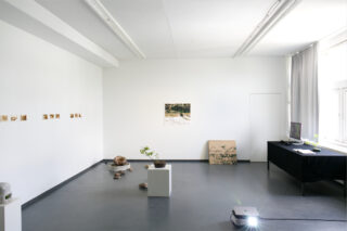 Eine Galerie für minimalistische Kunst mit weißen Wänden, Fotografien, kleinen Skulpturen auf dem Boden und auf Sockeln, einem Tisch mit einem Monitor sowie einem Projektor, der Licht auf den Boden wirft. Durch die Fenster fällt Tageslicht herein.