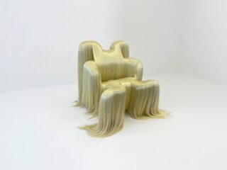 Ein beiger Sessel, dessen Oberflächen mit langem, glattem, blondem, haarähnlichem Material drapiert sind, steht in einem minimalistisch eingerichteten weißen Raum.
