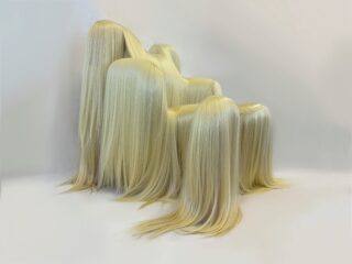 Vier abstrakte, kopfähnliche Objekte, die mit langen, glatten, blonden Haaren bedeckt sind, stehen dicht beieinander vor einem schlichten weißen Hintergrund.